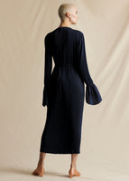 A model standing backwards wearing a navy blue pleated midi dress with thin tie at the waist.