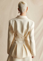 A model standing backwards wearing an ivory jacket with pleats in the back and a fabric belt. Buttons on cuff.