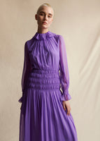 Image of a model wearing a violet long sleeved dress with smocking at the waist.