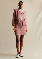 A model standing facing forward, wearing a pink and white tweed knit jacket with pearl buttons, open over a red and white striped shirt. She is wearing a matching pink and white tweed knit mini skirt that also has pearl buttons. 