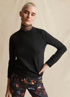 Model wearing a black crewneck sweater styled over a lace turtleneck. Wearing floral black pants.
