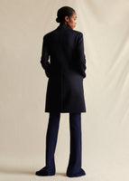Model standing backwards wearing a navy blue mid length coat, over navy blue flare pants.