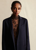 Image from the waist up of a model wearing a navy blue coat styled over a black lace turtleneck.