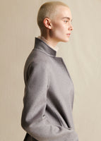 An image of a model standing sideways, wearing a grey cashmere coat. Image is from the waist up. 