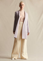 A model wearing a lilac mid-length coat, layered over an ivory lace turtleneck.  Styled with ivory wide leg pants.