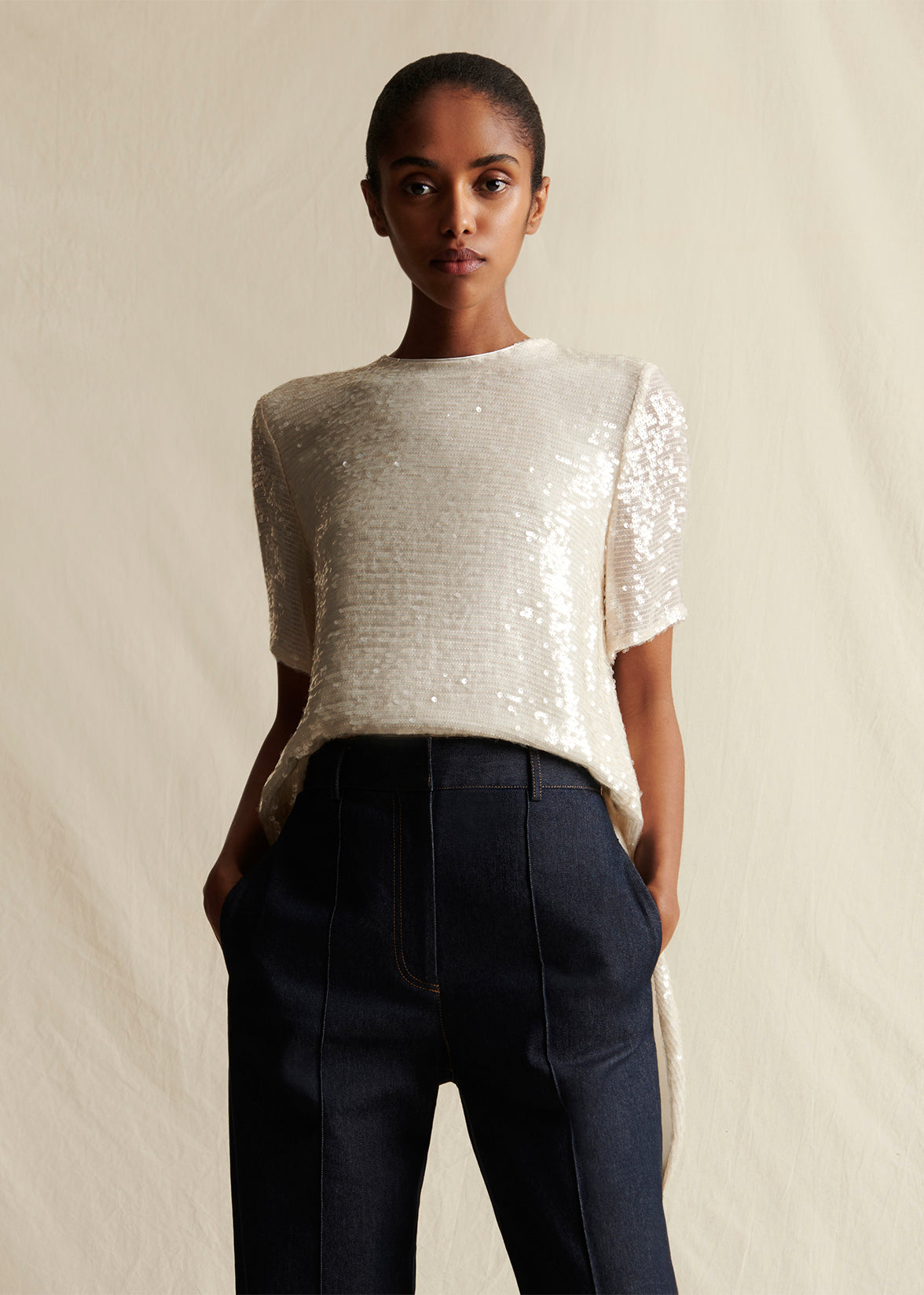 Model facing forwards wearing an ivory sequin short sleeve top with dark denim jeans by Adam Lippes