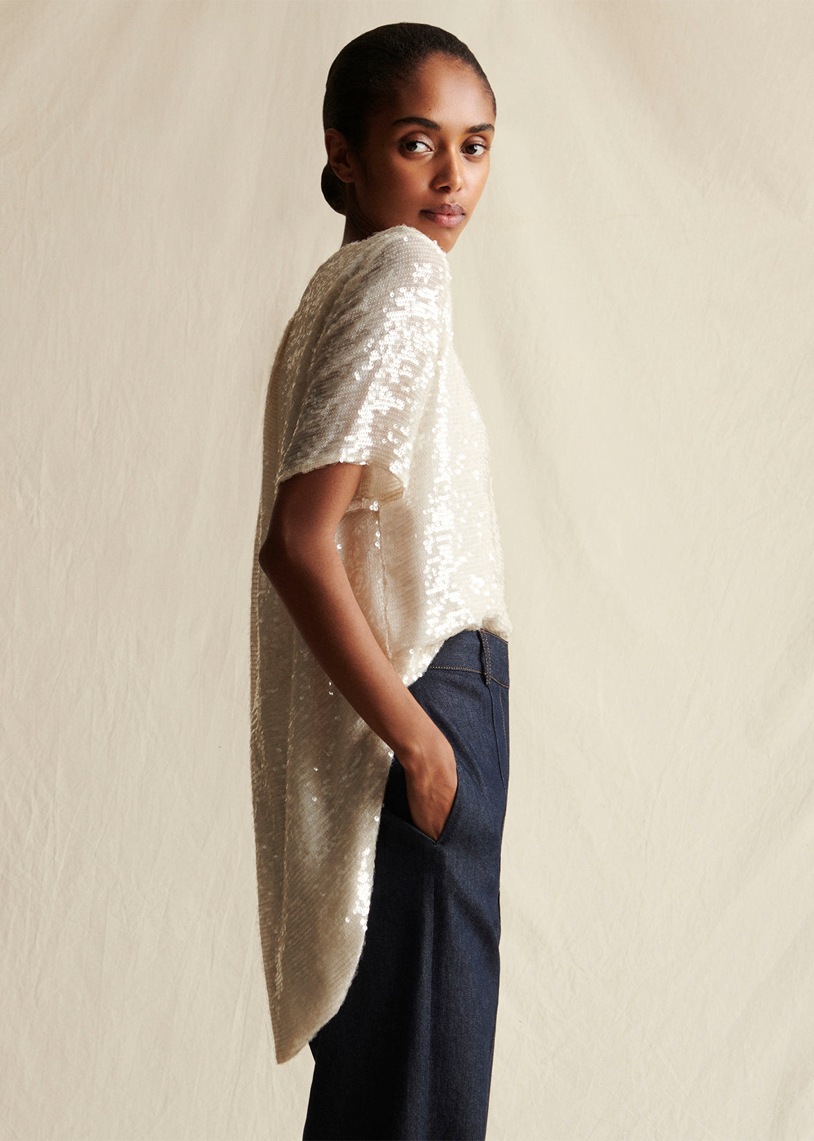 Model facing sideways wearing an ivory sequin short sleeved top and dark blue jeans by Adam Lippes