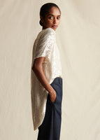 Model facing sideways wearing an ivory sequin short sleeved top and dark blue jeans.