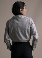 A back-facing image of a model wearing a long sleeve blouse with high neckline in stripe shirting with a bow tied at the front neck worn with black pants.