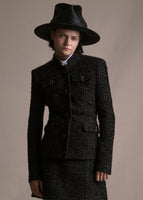 Model wearing mid length metallic jacket with pockets and crystal buttons across the front with matching skirt and a black hat.
