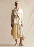 Image of a model wearing an ivory double breasted blazer with a waist tie. Paired with an ivory pleated mid-length skirt.