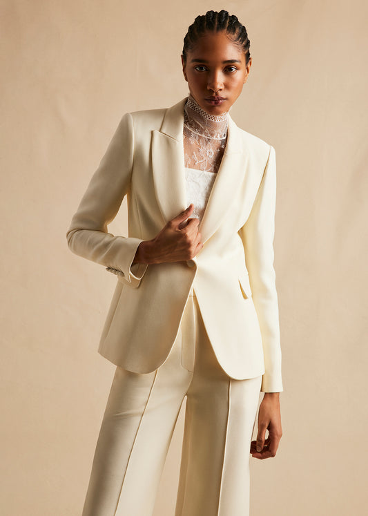 Image of a model facing forward wearing single breasted blazer in silk wool in ivory over an ivory lace turlteneck. With ivory pants.