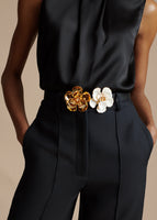Cropped image of a model wearing a black silk top with black pants. She is also wearing a leather belt with two large metal flowers on it - one gold flower, one white flower.