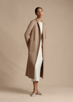 An image of a model wearing a camel color long coat over a white tea length dress.