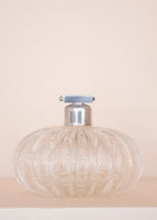A glass perfume bottle with a silver closure on a pale pink background. 
