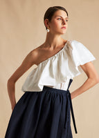 A model wearing a white poplin one shoulder top tucked into a navy wrap skirt with a pearl earring.