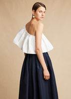 A side view image of a model wearing a white poplin one shoulder top tucked into a navy wrap skirt with a pearl earring.