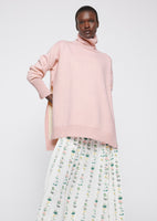 A model wearing a pink turtleneck sweater over a pleated white floral dress.